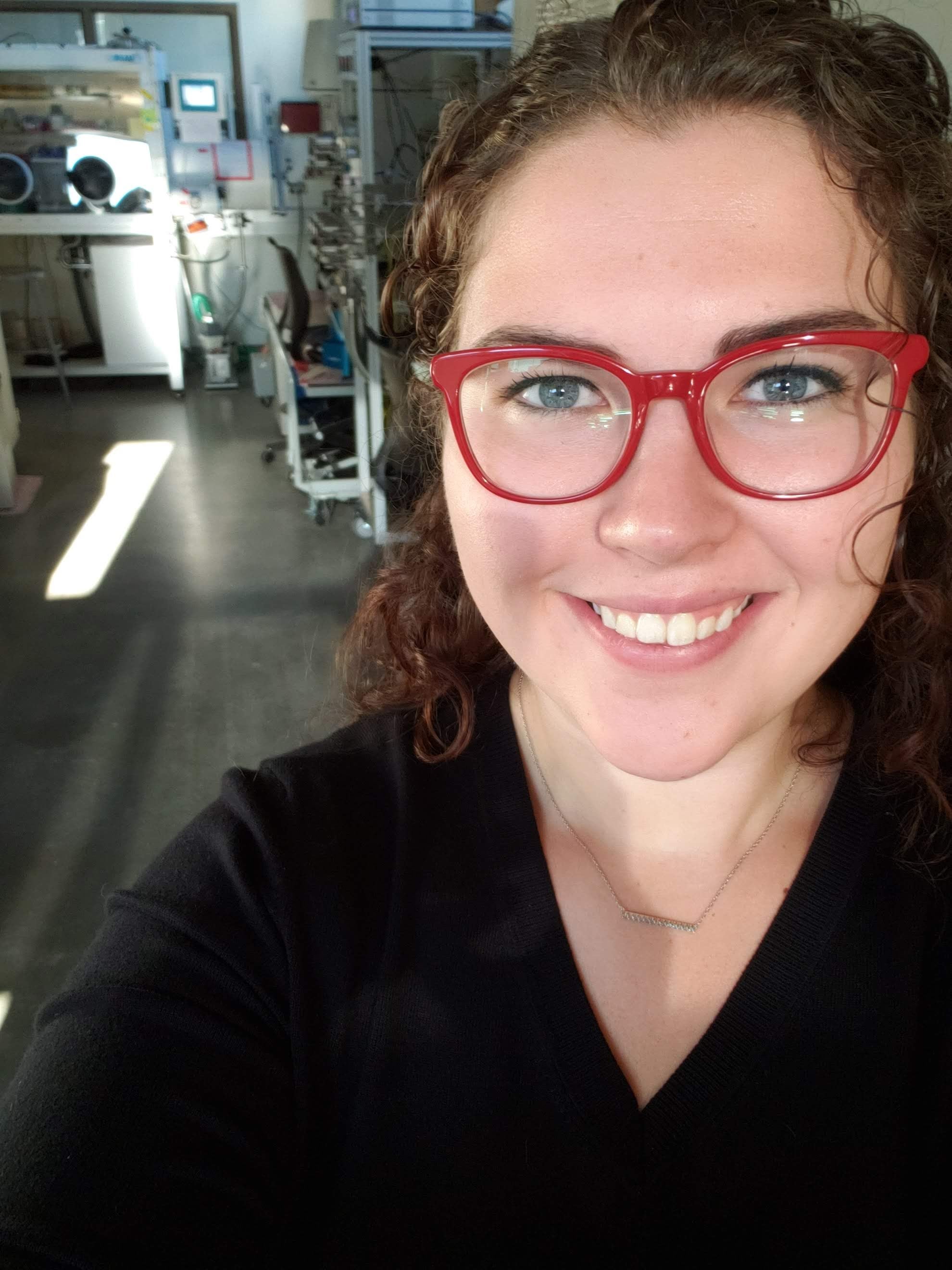 Selfie of Erin Taylor, wearing red framed eyeglasses, with lab equipment in the background