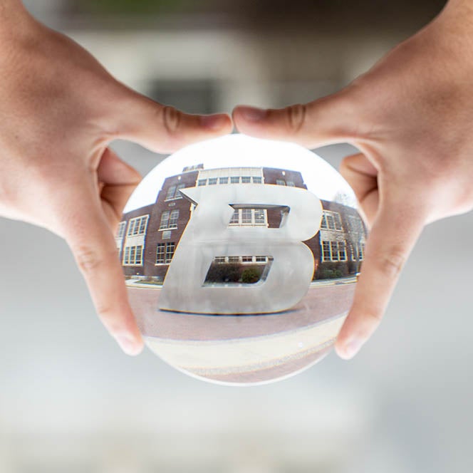 B statue reflected in a glass orb, held by two hands