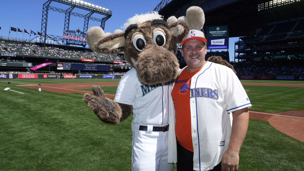 A person in a Mariners jersey and cap smiles with a moose mascot on a baseball field