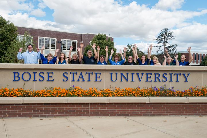 A group of employees posing and waving behind a Boise State University sign