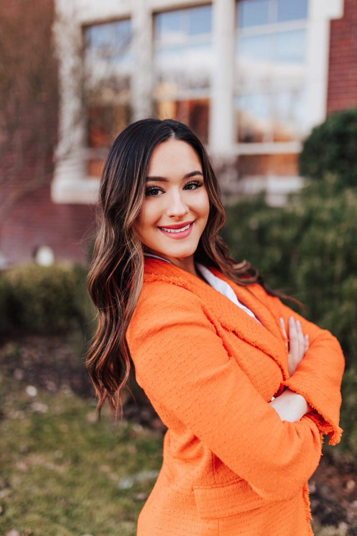 A woman in an orange blazer folds her arms and smiles in front of a brick building with a prominent glass window