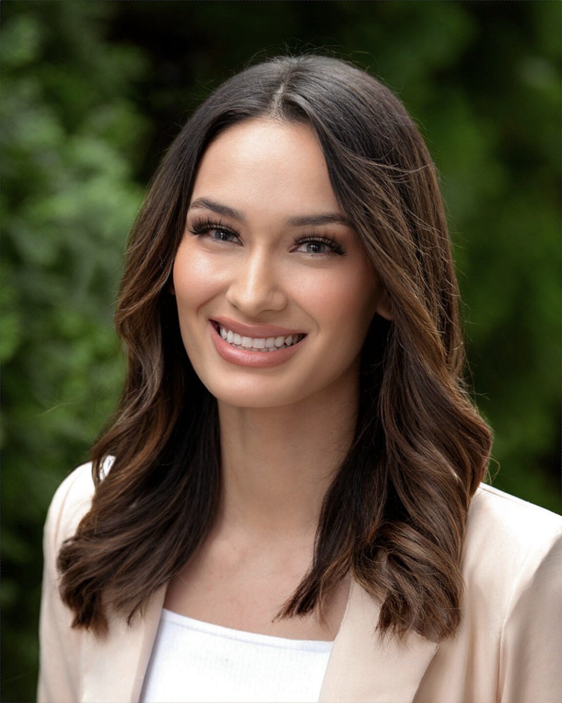 A woman with brown hair and a tan blazer smiles in front of a green background