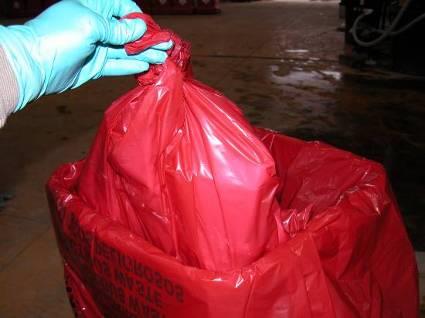 photo of a gloved hand depositing a red Regulated Medical Waste bag with balloon style knot into an approved receptacle