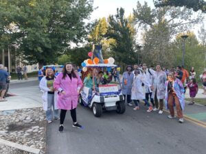 Photo of the Chemistry Club members wearing lab coats and standing next to their parade float made with balloons on a golf cart.