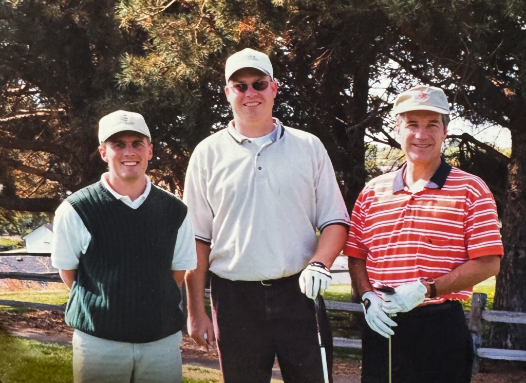 Three men in golf attire stand on a course with trees and a fence in the background