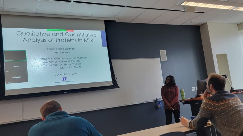 Photo of Rianat Lukman standing next to her presentation's title slide, "Qualitative and Quantitative Analysis of Proteins in Milk"