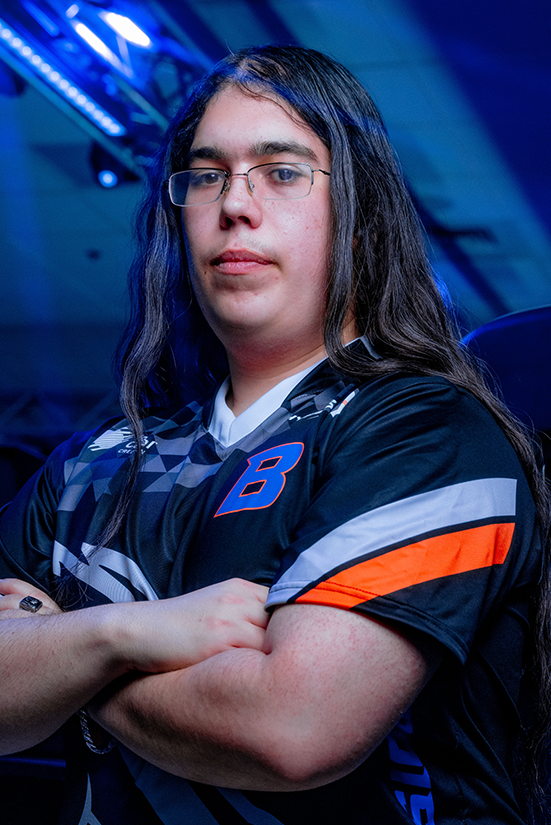 A collegiate esports player sits in a dimly lit modern esports arena, looking at the camera, arms folded.