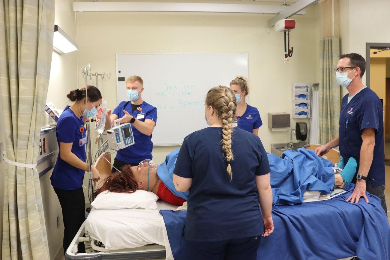 Students in scrubs stand around a large manikin lying on a hospital bed.