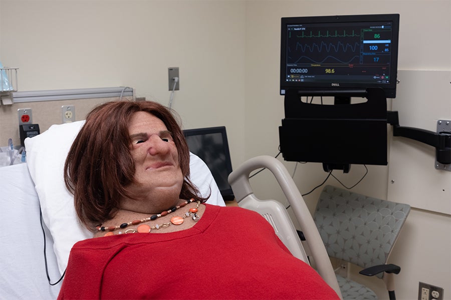 Carly the manikin sits in a hospital bed next to a screen displaying vital signs