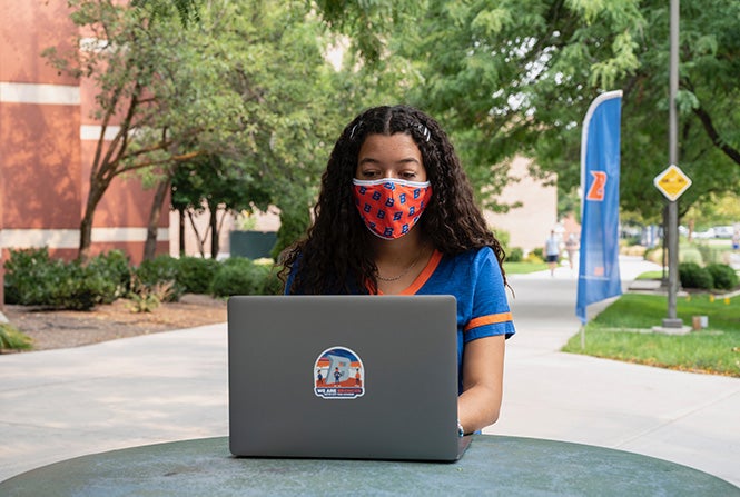 Student wearing facial covering sits outside on campus and works on laptop