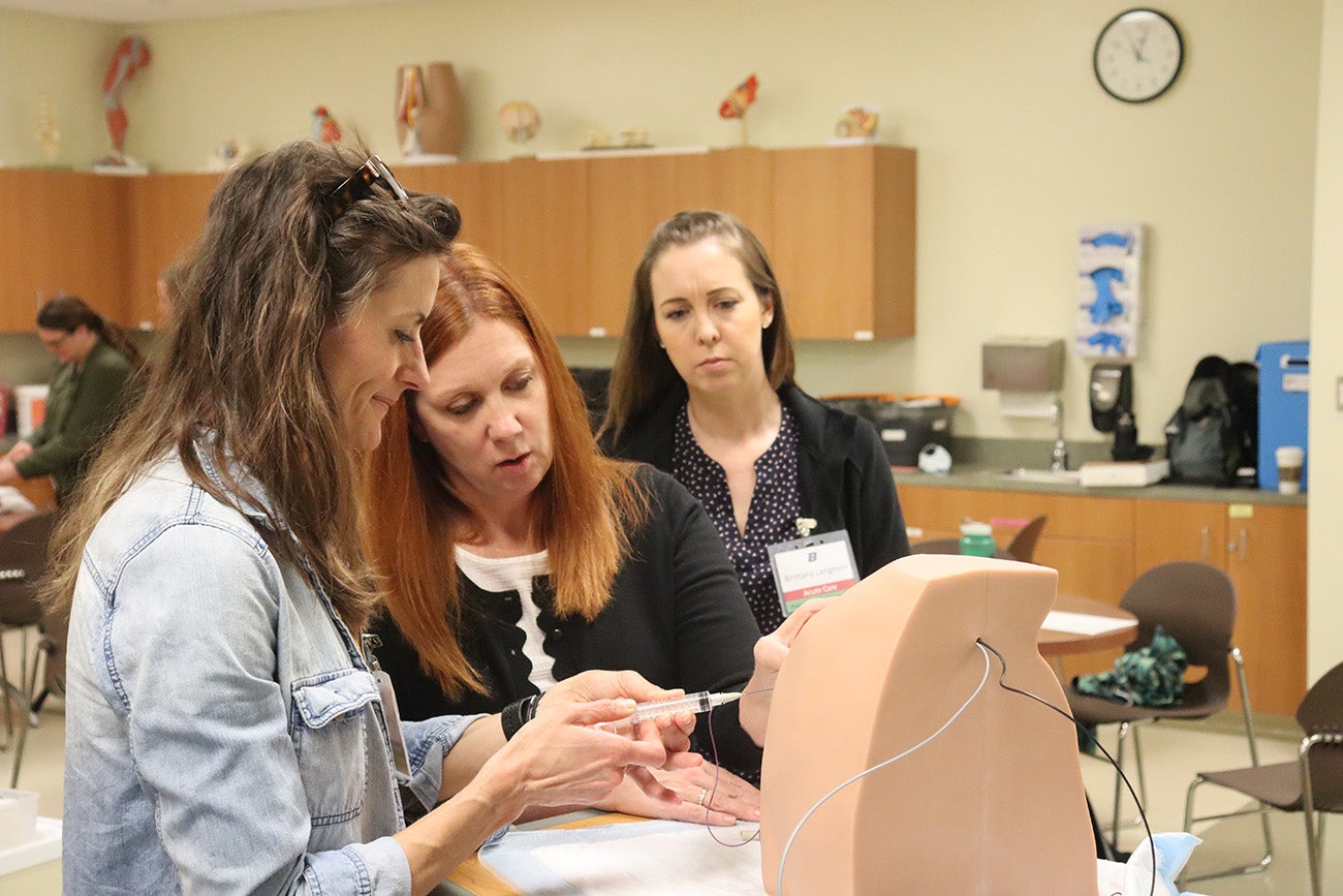 A student in the foreground inserts a needle into a manikin as a nurse practitioner professor guides her. Another student looks on.