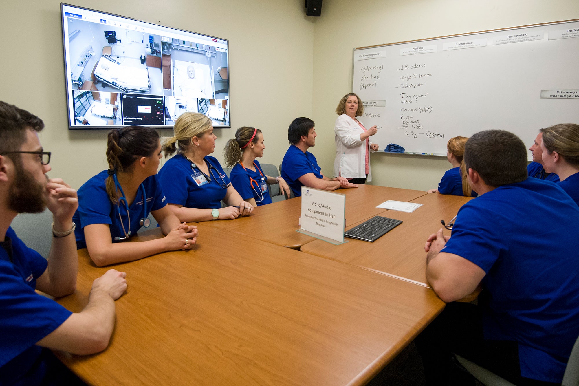 A professor writes on a whiteboard at the front of a room while a group of students in blue scrubs sit around a conference table.