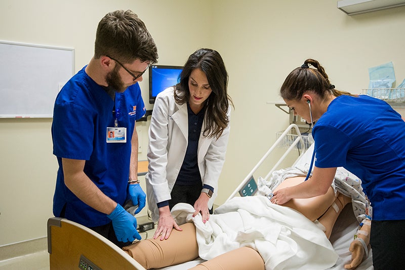 A professor in a white coat shows something on a manikin's leg to a student. Another student uses a stethoscope to listen to the manikin's stomach.