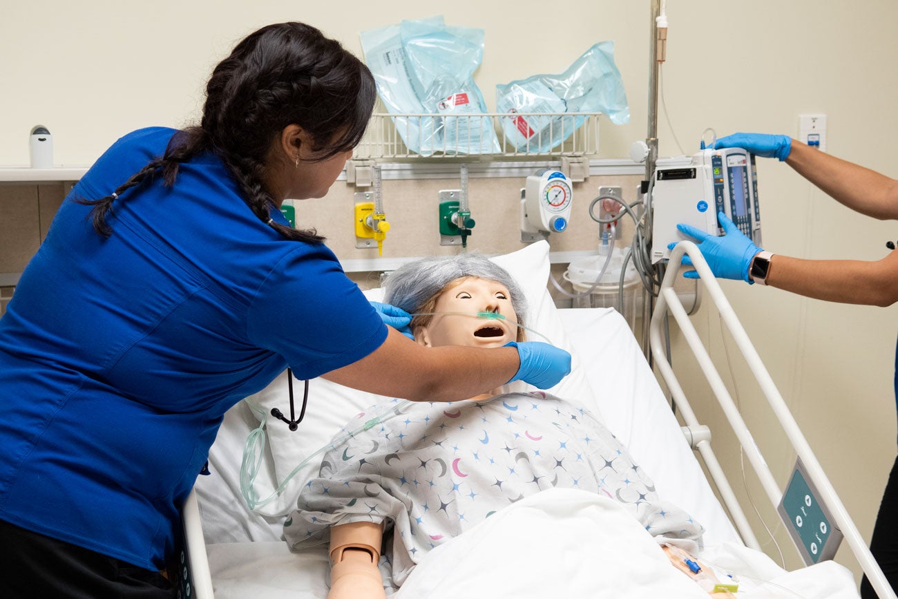 A nursing student adjusts an oxygen tube on a manikin wearing a gray wig.