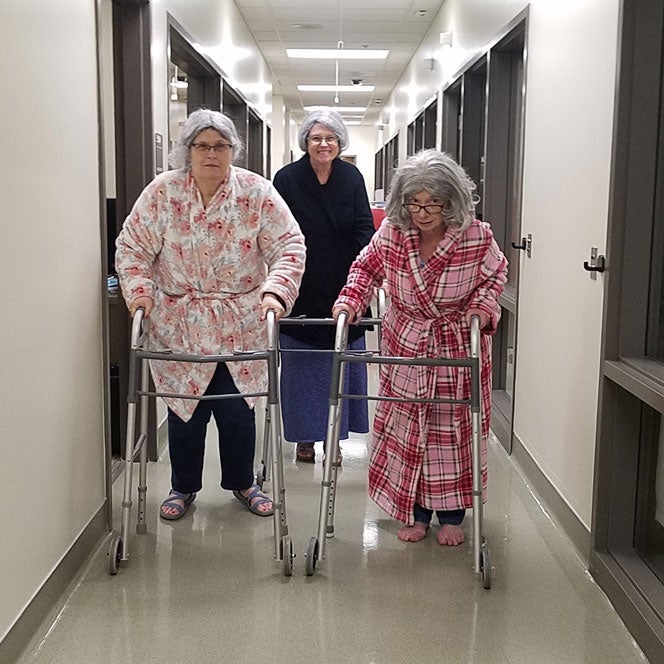 There elderly women wearing bathrobes push walkers down the simulation center hallway.