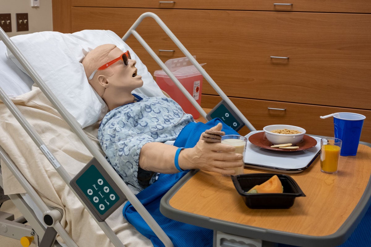 A Hal manikin eats breakfast while lying in a hospital bed.
