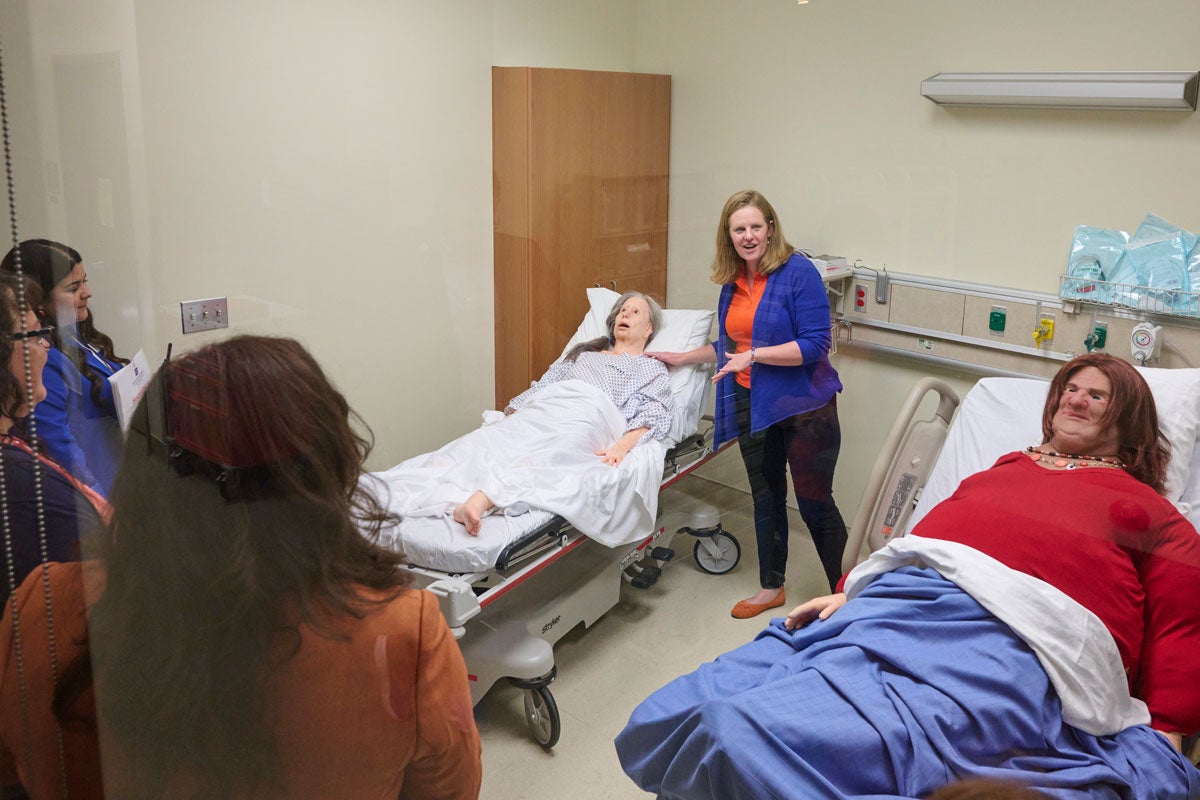 A professor stands between a geriatric manikin and a bariatric manikin lying on hospital beds.