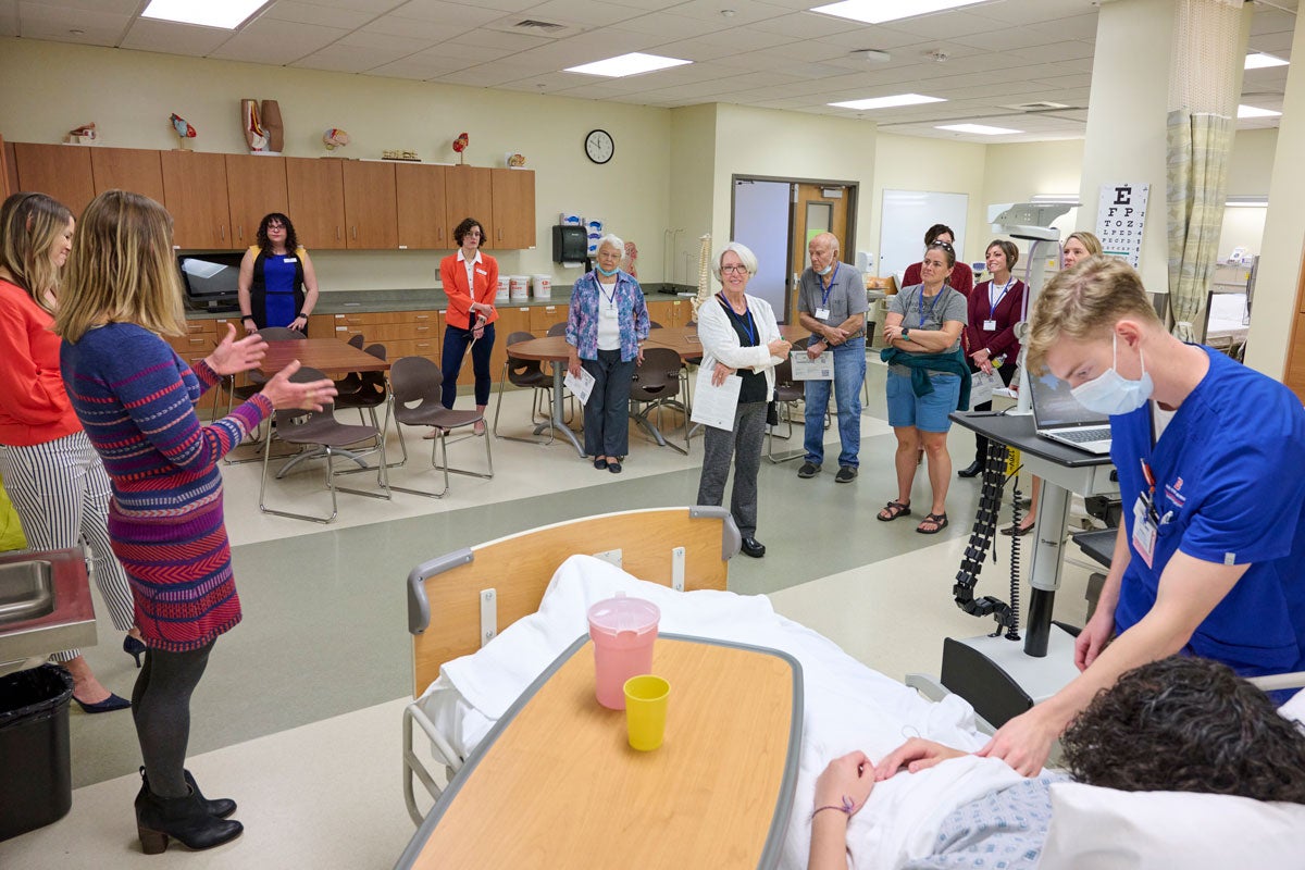 In the foreground, a student nurse tends to a standardized patient. In the background, a professor talks to a tour group.