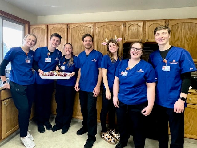 Seven nursing students stand in a kitchen, smiling in their blue scrubs and holding a plate of veggie skewers.