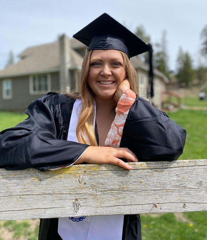 Libby Sheffler leans on a wood fence rail in her Lewis-Clark State College regalia