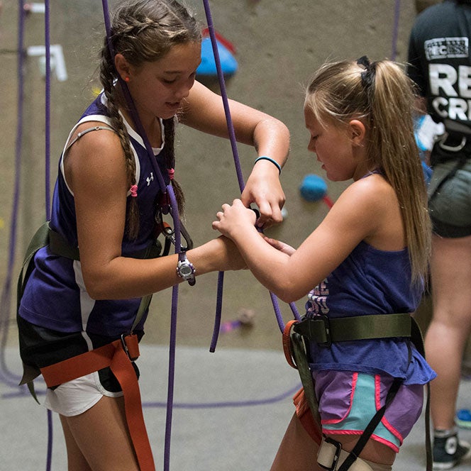 Two young female children adjust their harnesses and ropes in front of a rock climbing wall