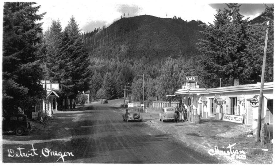 Black and white phote of gas station surrounded by mountains and pine trees, cars driving on dirt road in front of the station
