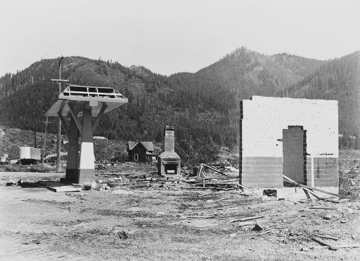 black and white photo showing remains of station, only a brick wall and chimney remain of building