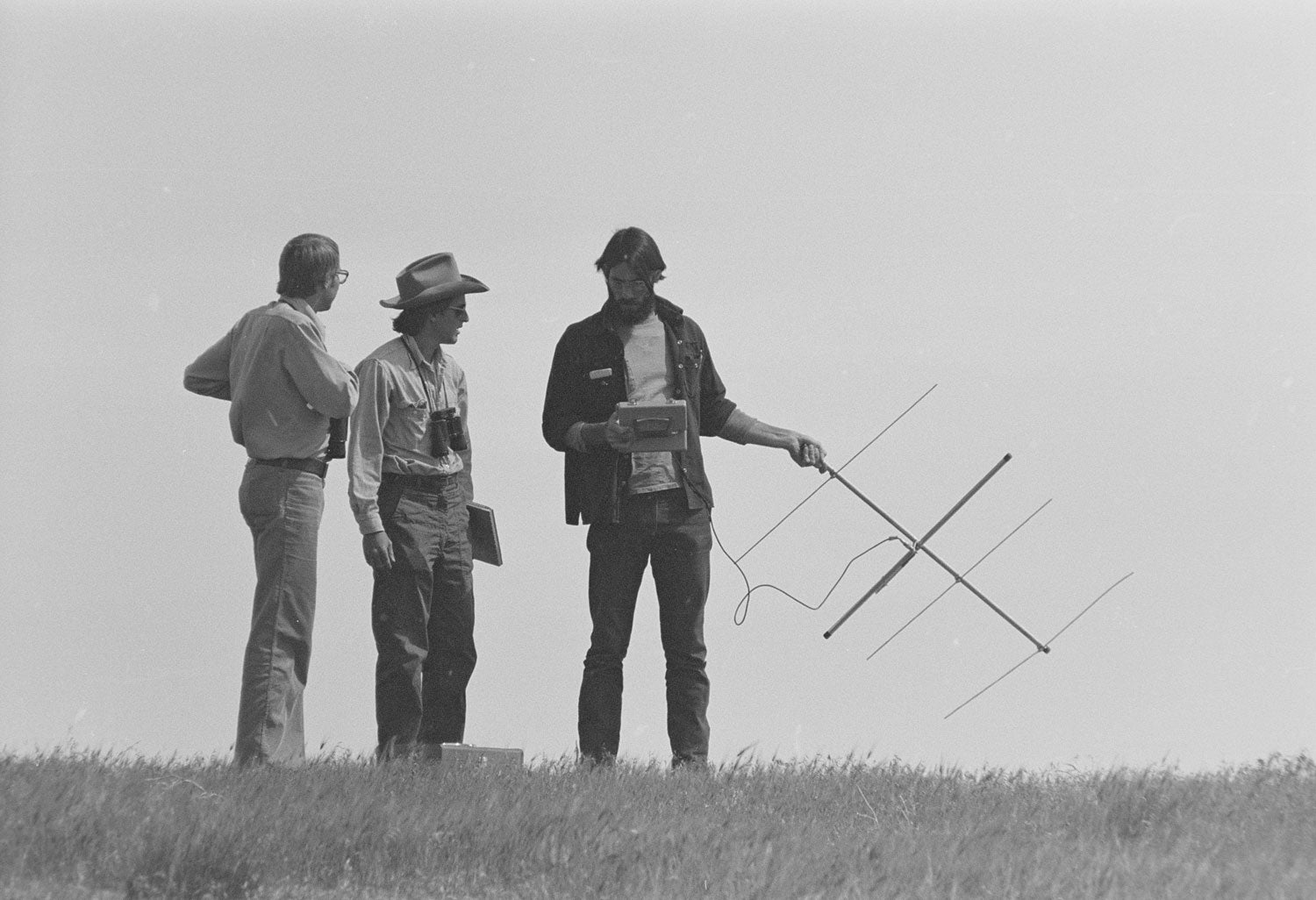 a black and white photo shows three men. One is holding an antenna and has long brown hair and a beard styled in a 1970s fashion. the second man has a large cowboy hat and aviator sunglasses. The third man has long 1970s style hair and large-lensed glasses