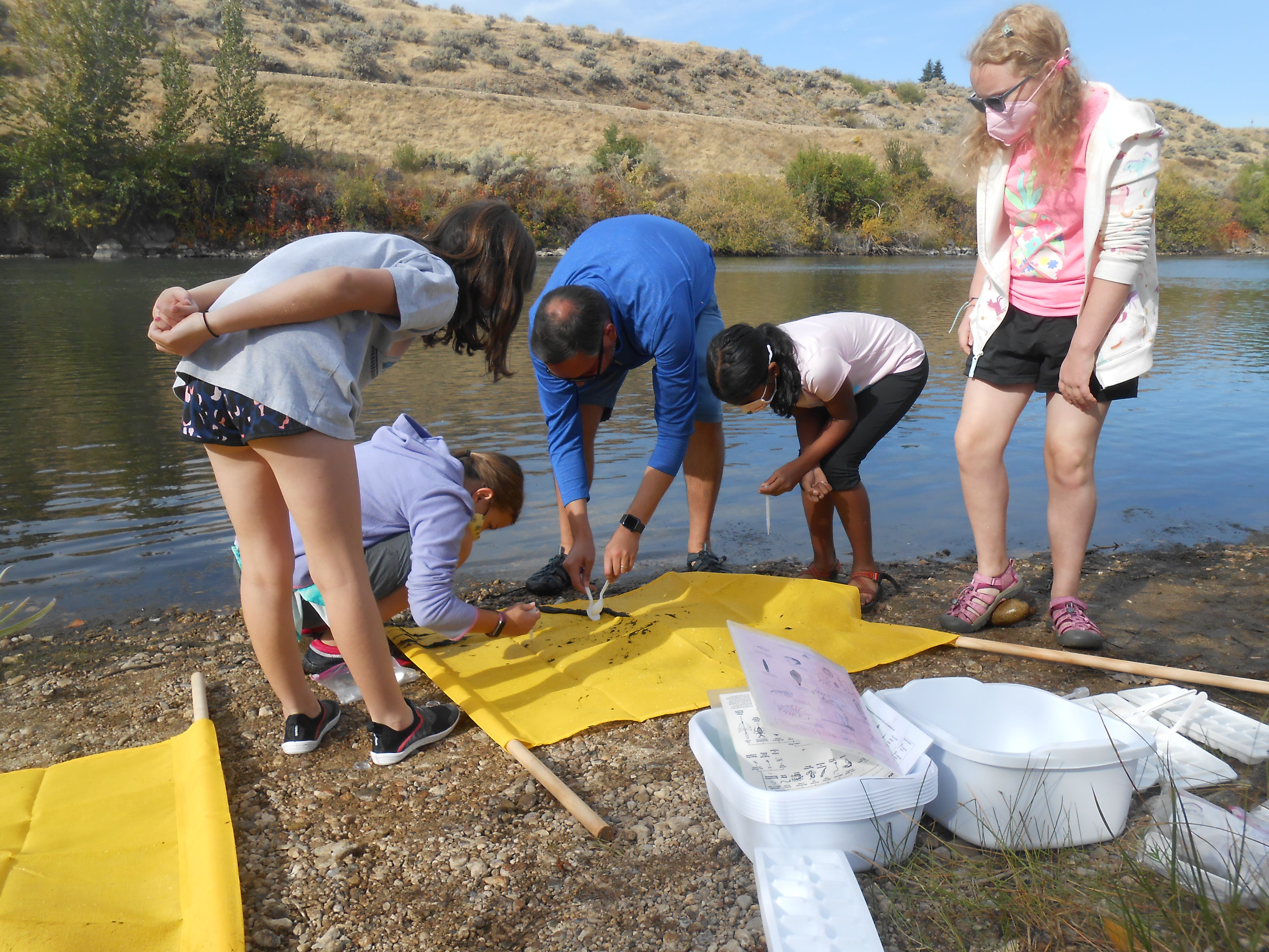 a teacher and four students stand on the river bank. They stoop to look closely at a yellow tarp that has mud and debris spread on it