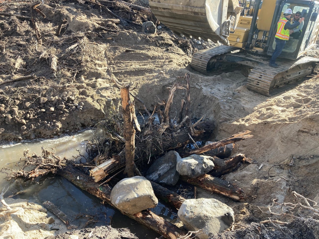 Large tree trunks with root wads are shoved into the mud along the side channel bank, with large boulders stacked on top. A backhoe is visible in the background.