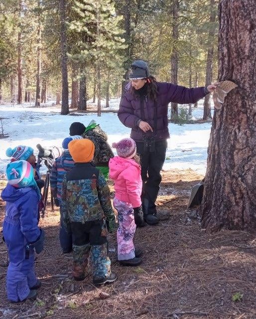 six small children stand looking at Heather as she holds out an owl wing against tree bark