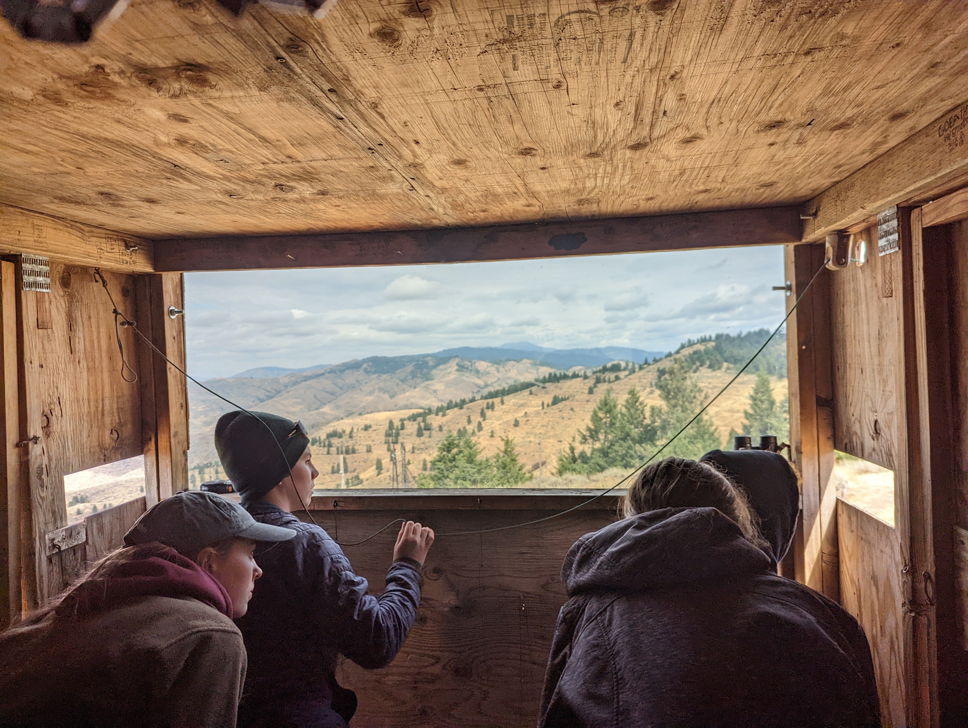 a view from inside the trapping blind looking out. Four people crouch inside a wooden shed, looking out the windows. Through the largest front window you can see a view of the Boise Ridge with mountains, trees, and grassy foothills