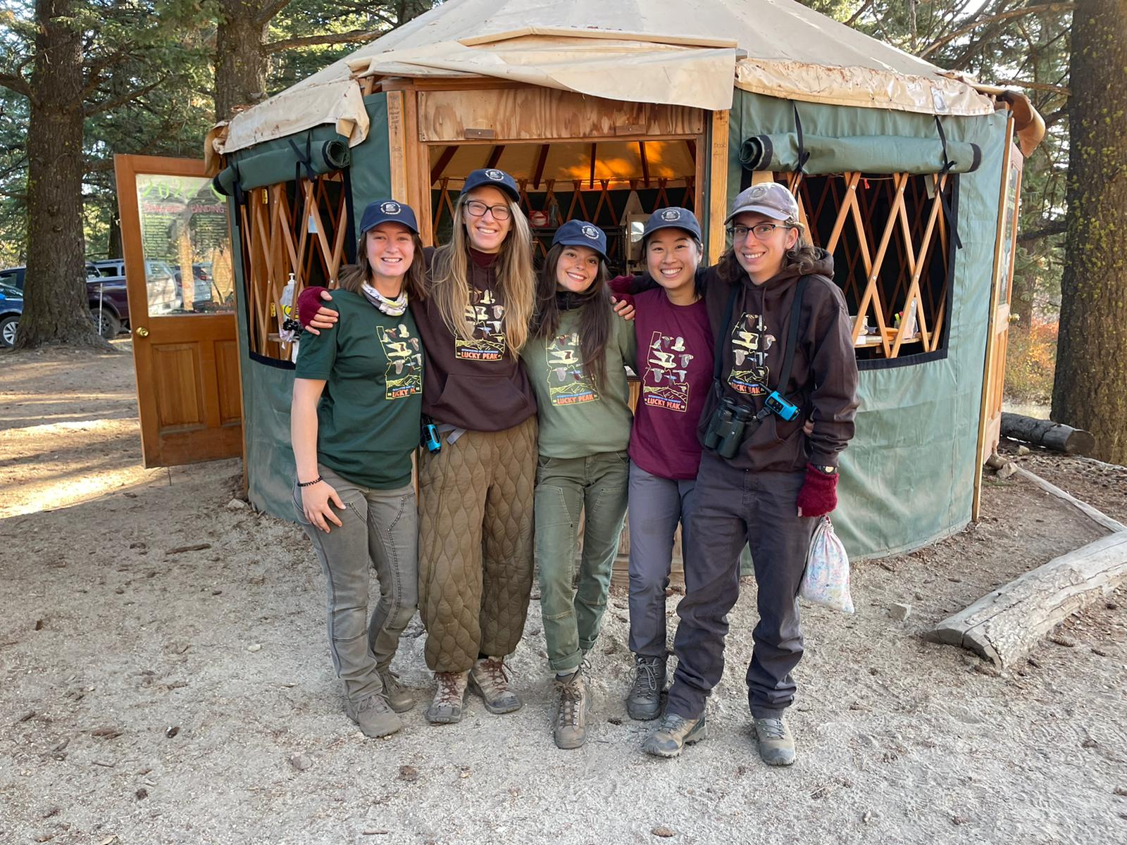 five biologists standing in front of a green yurt outside in the forest. They are smiling cheerfully and hugging each other, wearing matching IBO shirts and hats