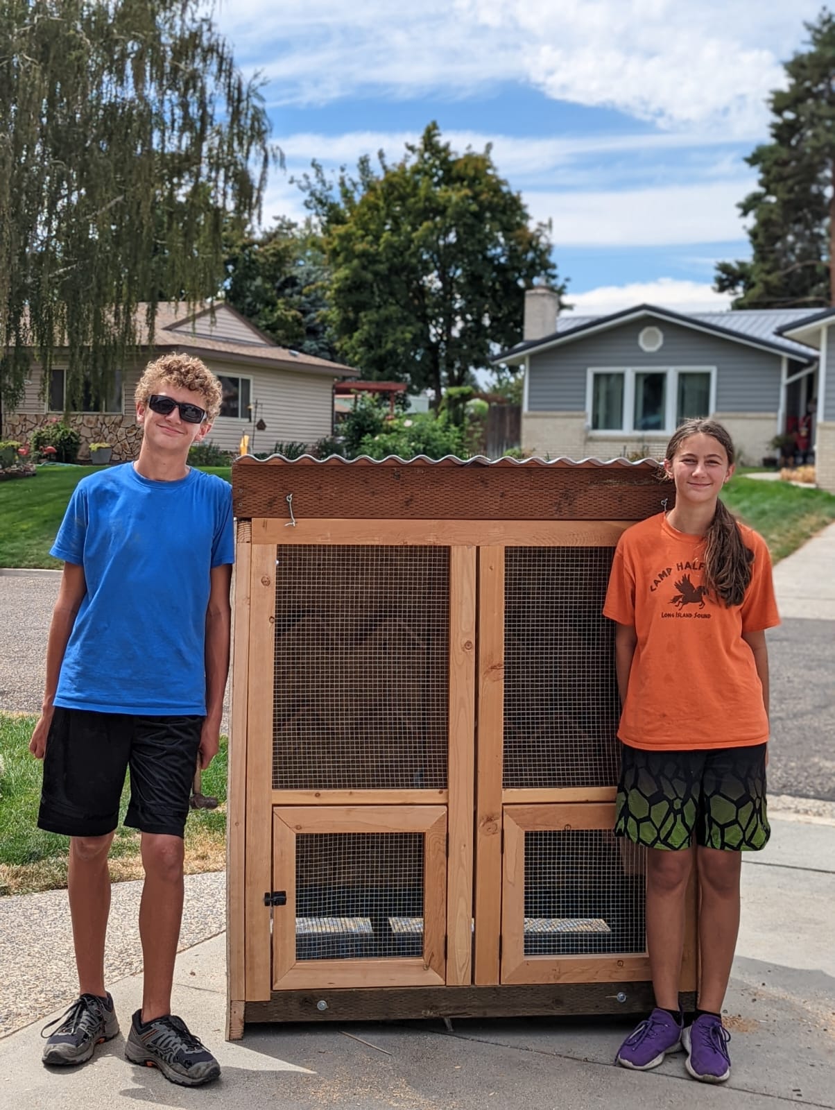two teens stand next to a small wooden structure that looks similar to a chicken coop or rabbit hutch