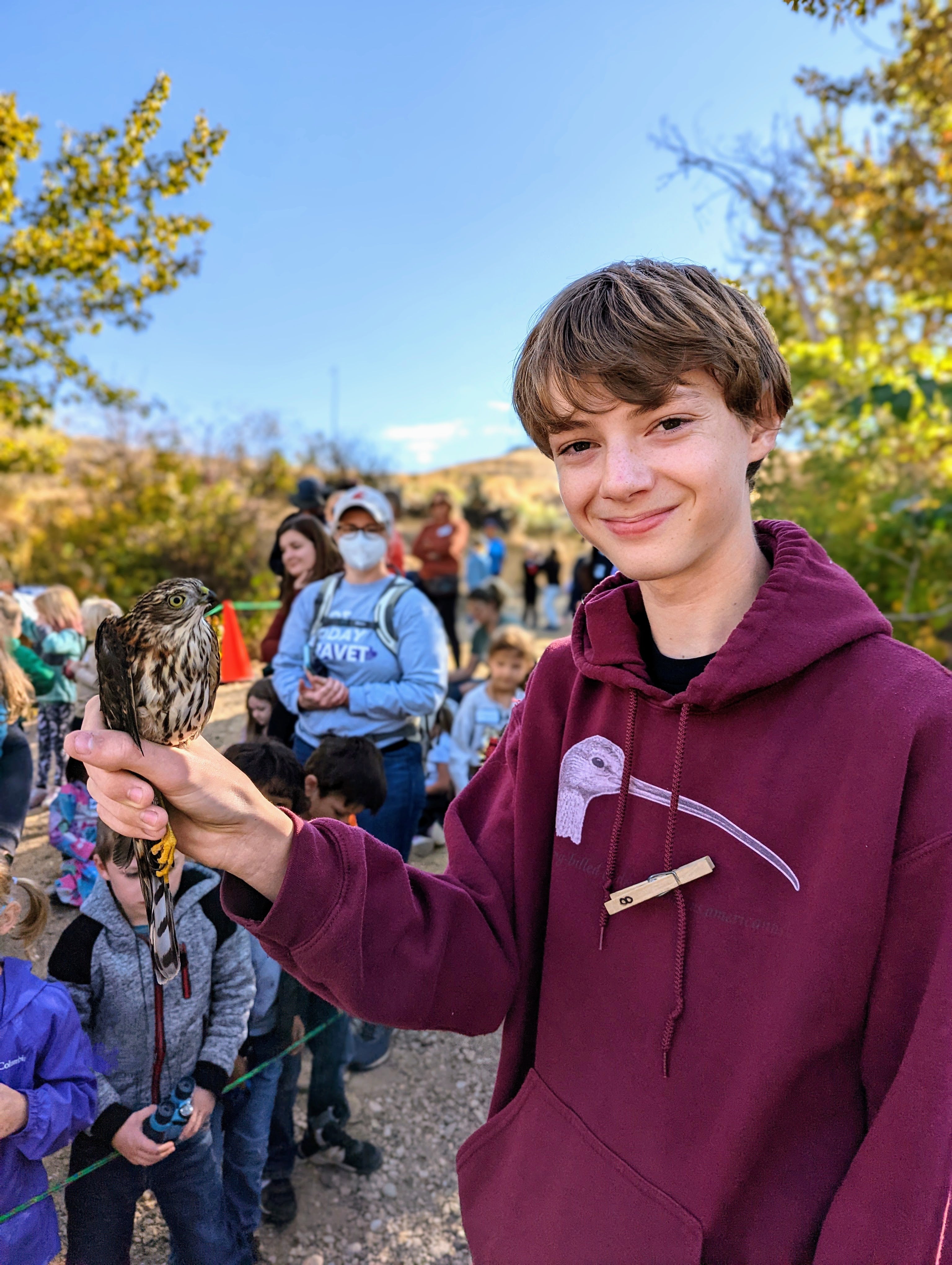 a high school student wearing an IBO sweatshirt is smiling while he holds a small brown streaked hawk. A crowd of small children and parents stands in the background