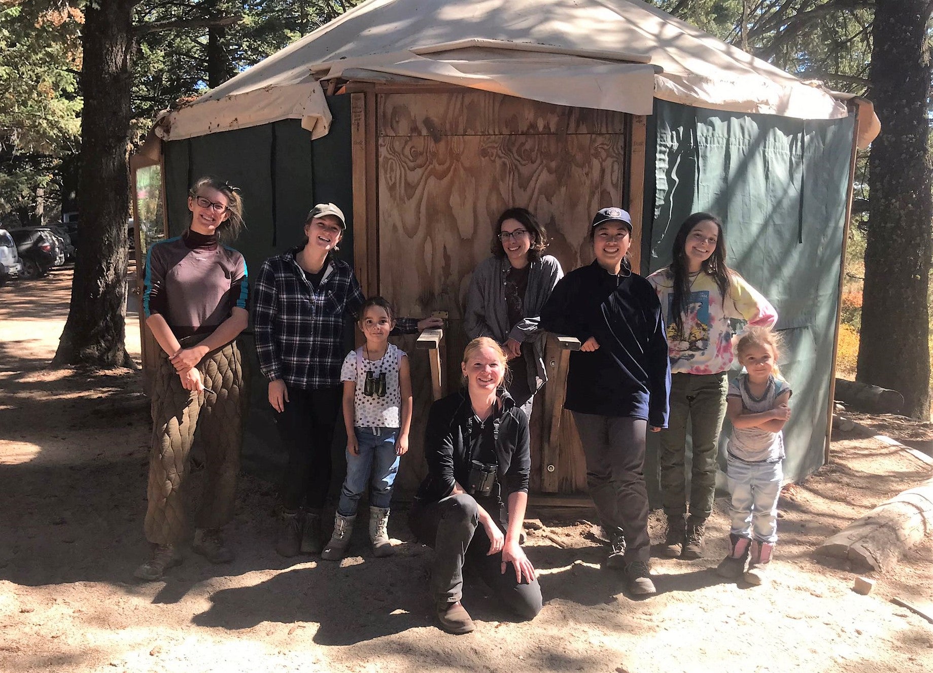 a group of scientists posing for a picture with smiling visitors outside in front of a green yurt under some trees