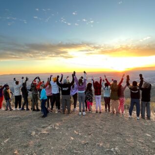 a group of about 20 students and a few parents stand at the top of Lucky Peak overlooking Boise. They are facing away over the valley, raising their arms into the air in celebration. The sun is setting with a beautiful orange glow