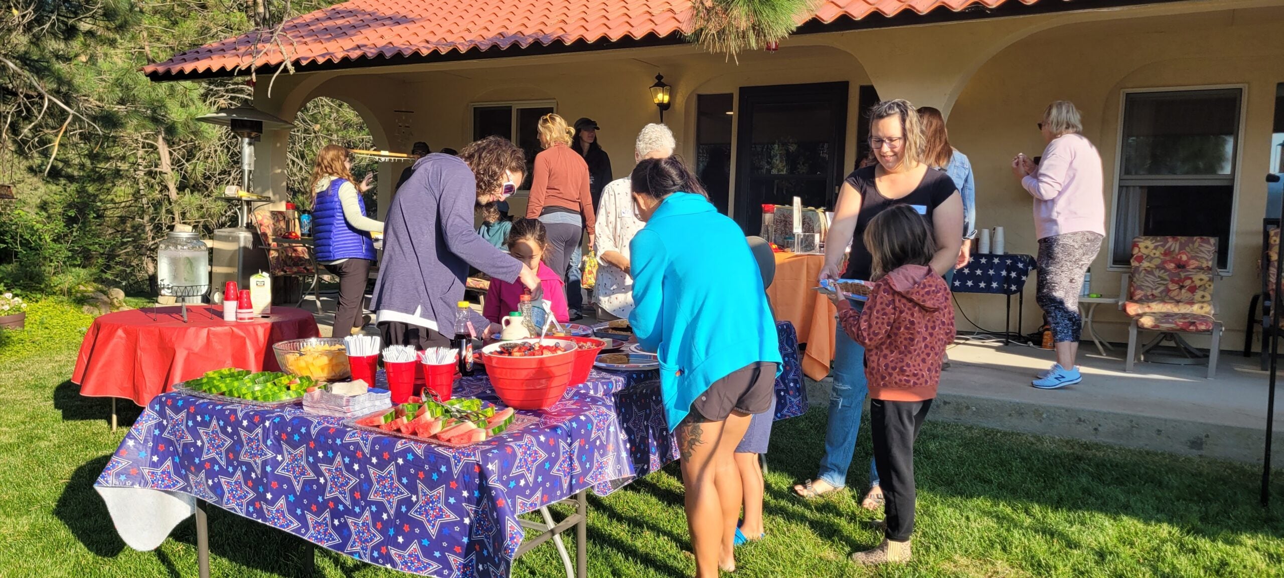 a group of people dish up food from tables spread with fruit, coffee, waffles, plates and bowls. the familiar view of Jennifer Alban's yard shows conifer trees, lawn, and and her home with red tile-style roof