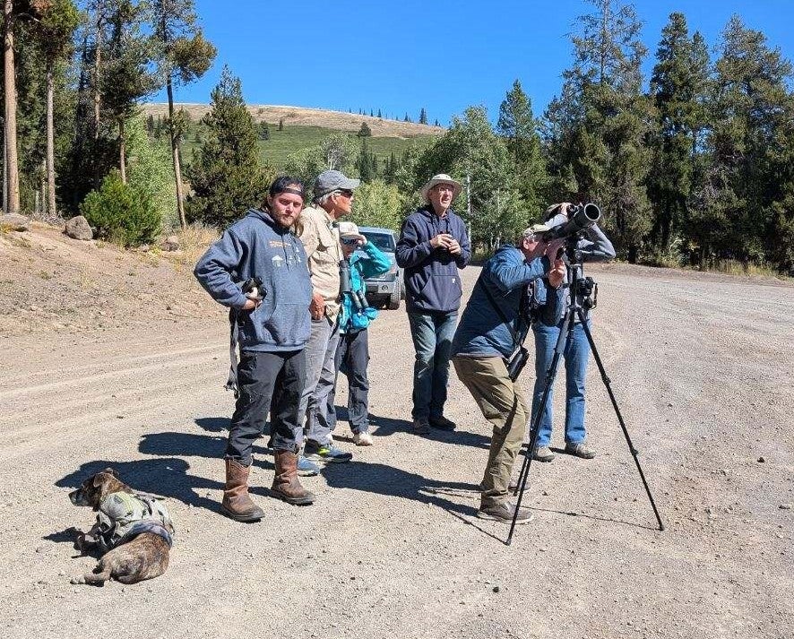 a group stands near a spotting scope. Some are looking through the scope, others binoculars. Ryan (left) stands looking at the camera wearing a hoodie and leather boots. Perl the dog lays near his feet looking relaxed and maybe a little bored