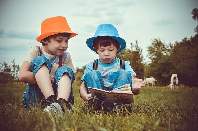 Color photo of two kids reading in a meadow and wearing colorful hats. Image by Artist and zabiyaka from Pixabay