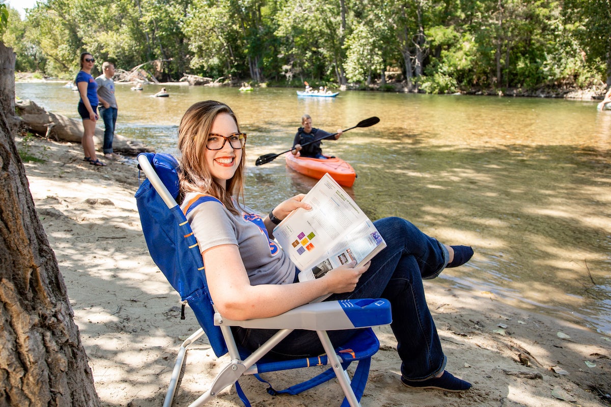 Student sits in beach chair, holding a text book, on the banks of the Boise River during a sunny summer day.
