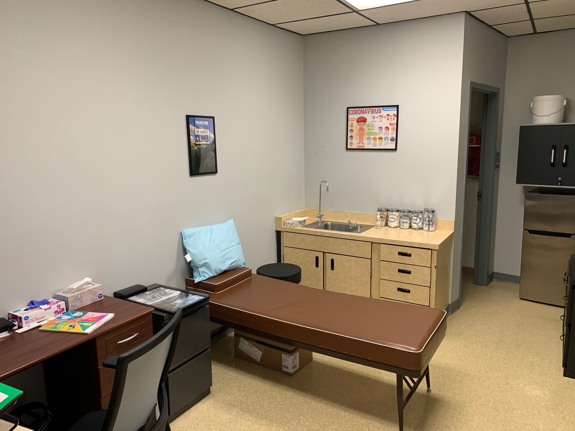 An exam room at the Marsing Elementary School clinic. The room include an exam bed, a desk and chair, and a sink and counter area with jars of tongue depressors, cotton swabs, and disinfectant wipes. 