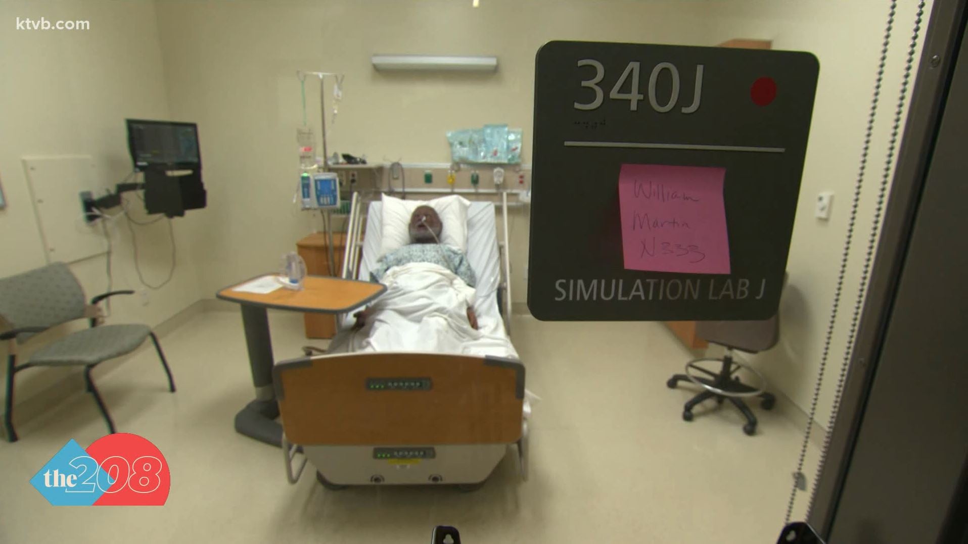 View through an observation window into a simulation hospital room. An adult male mannikin rests on the bed with a nasal tube inserted. A labelplate is mounted on the window indicating the room is a simulation lab. 
