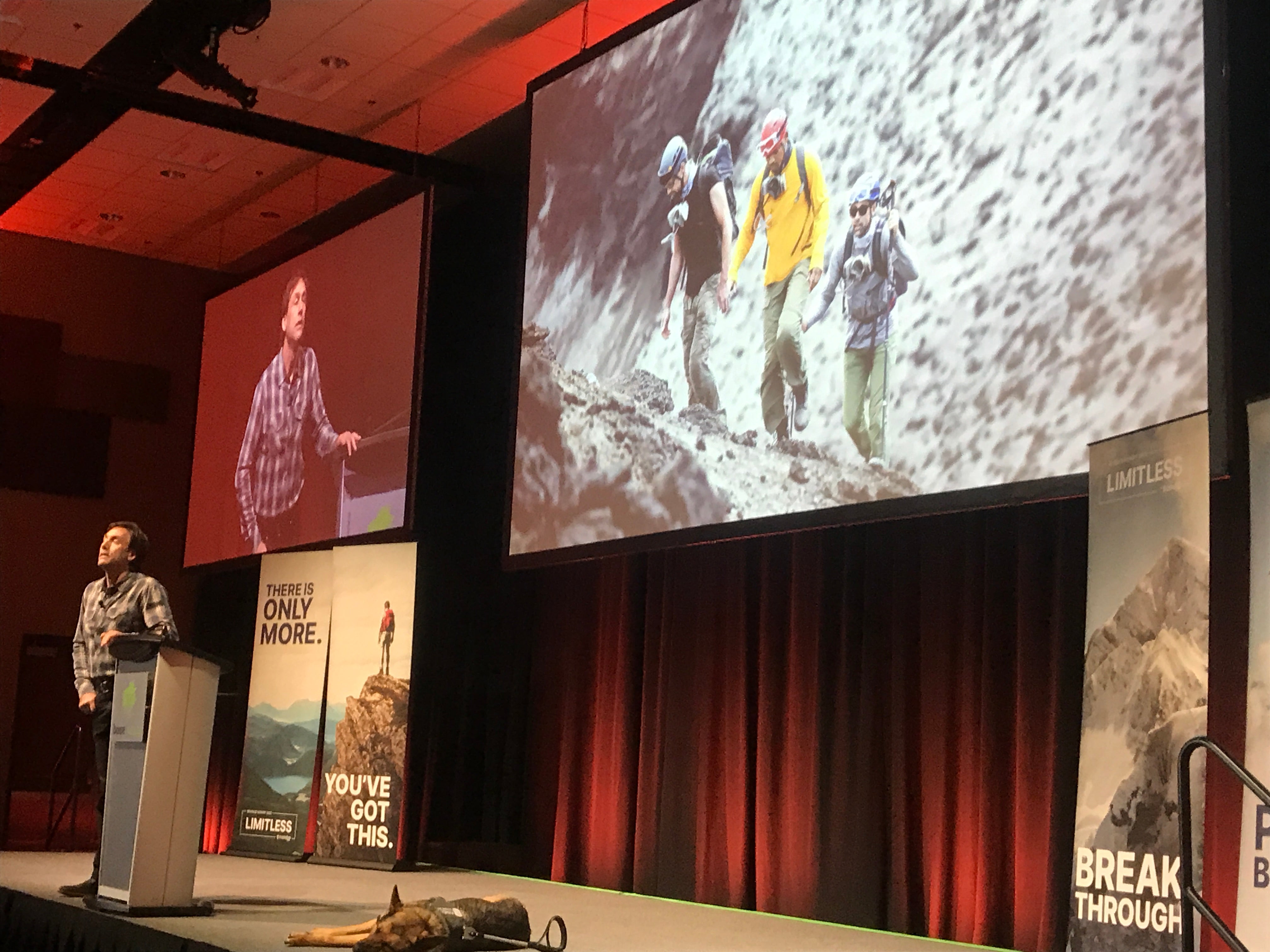 Erik on stage; service dog sleeps at his feet, presentation shows video of volcano trip