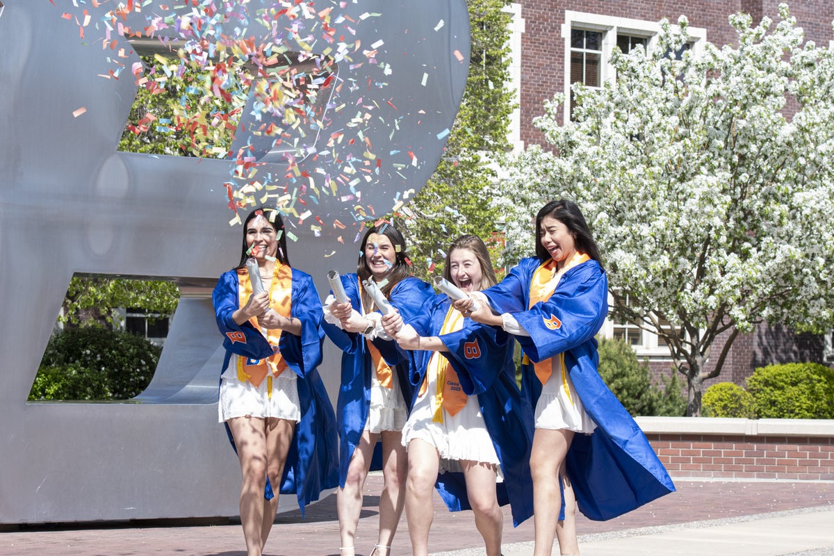 Four students in regalia pop confetti in front of the B statue 