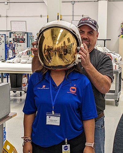 student in blue shirt wears gold screened astronaut helmet, man behinds her helps her put it on