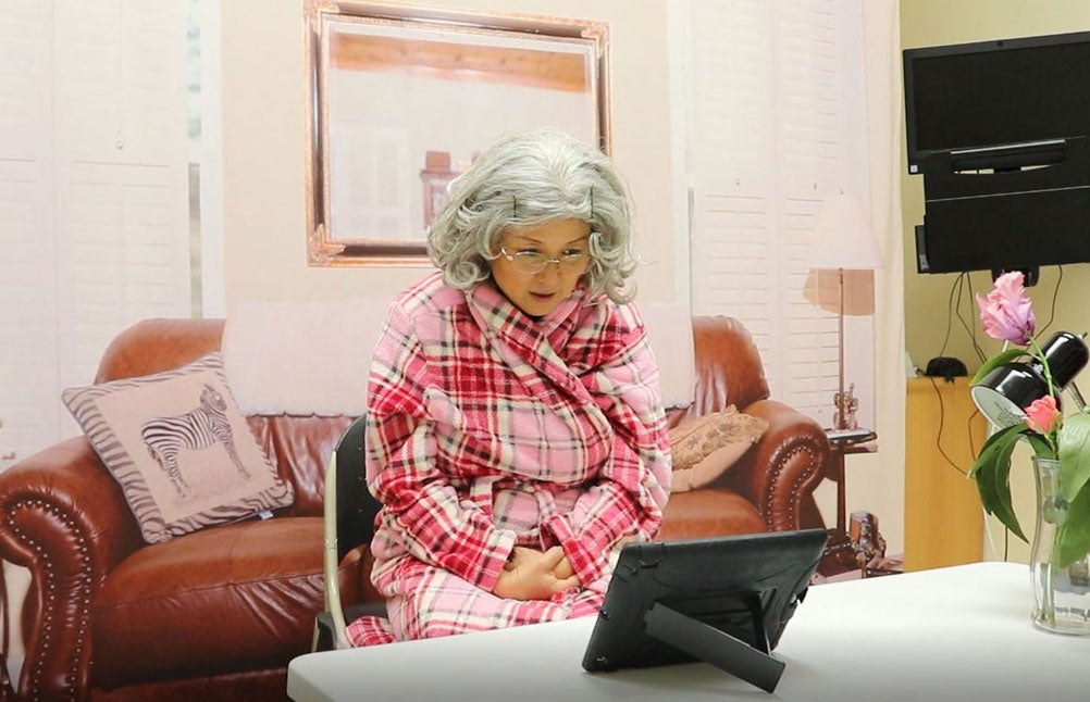 An elderly woman sits in a living room in the simulation center, looking at an electronic tablet. 