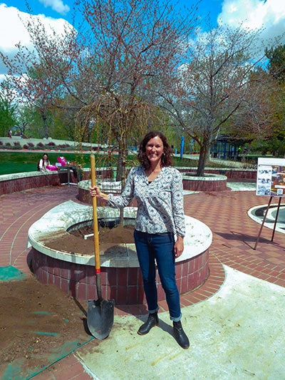 woman holds shovel in front of newly-planted tree
