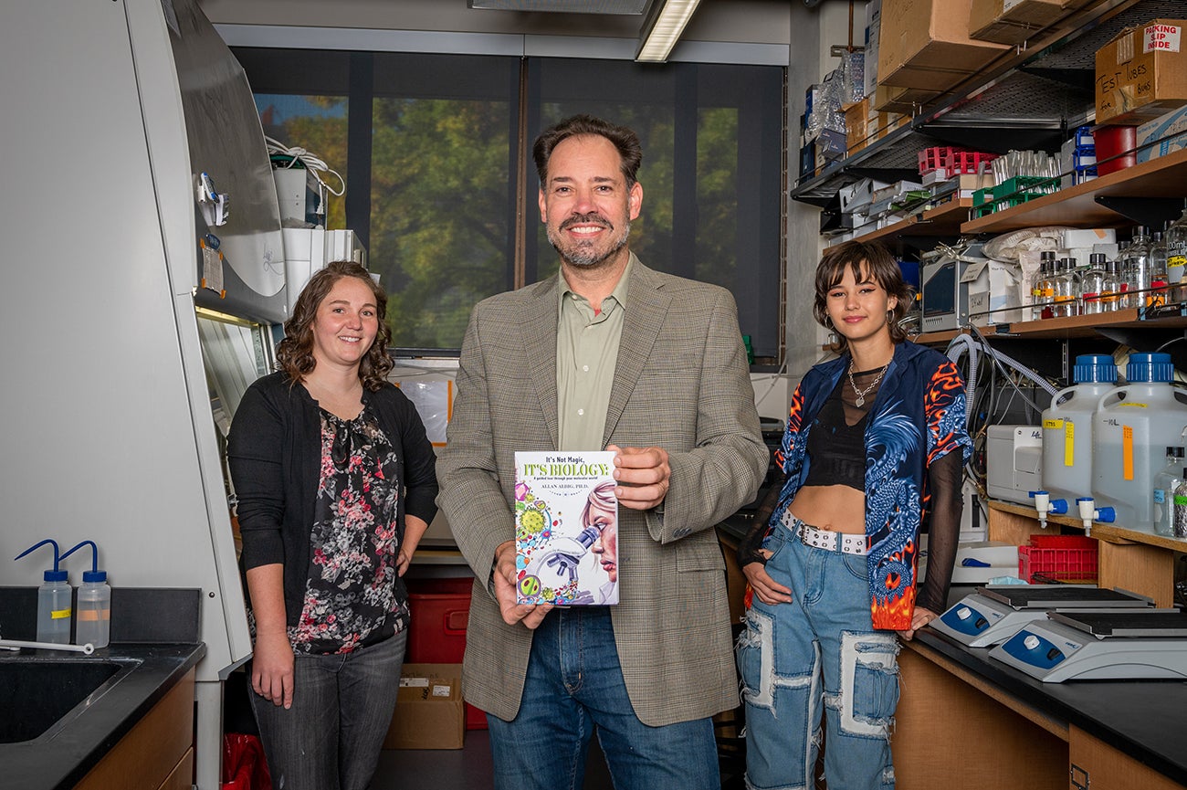 three people in caption stand in lab space holding self published book