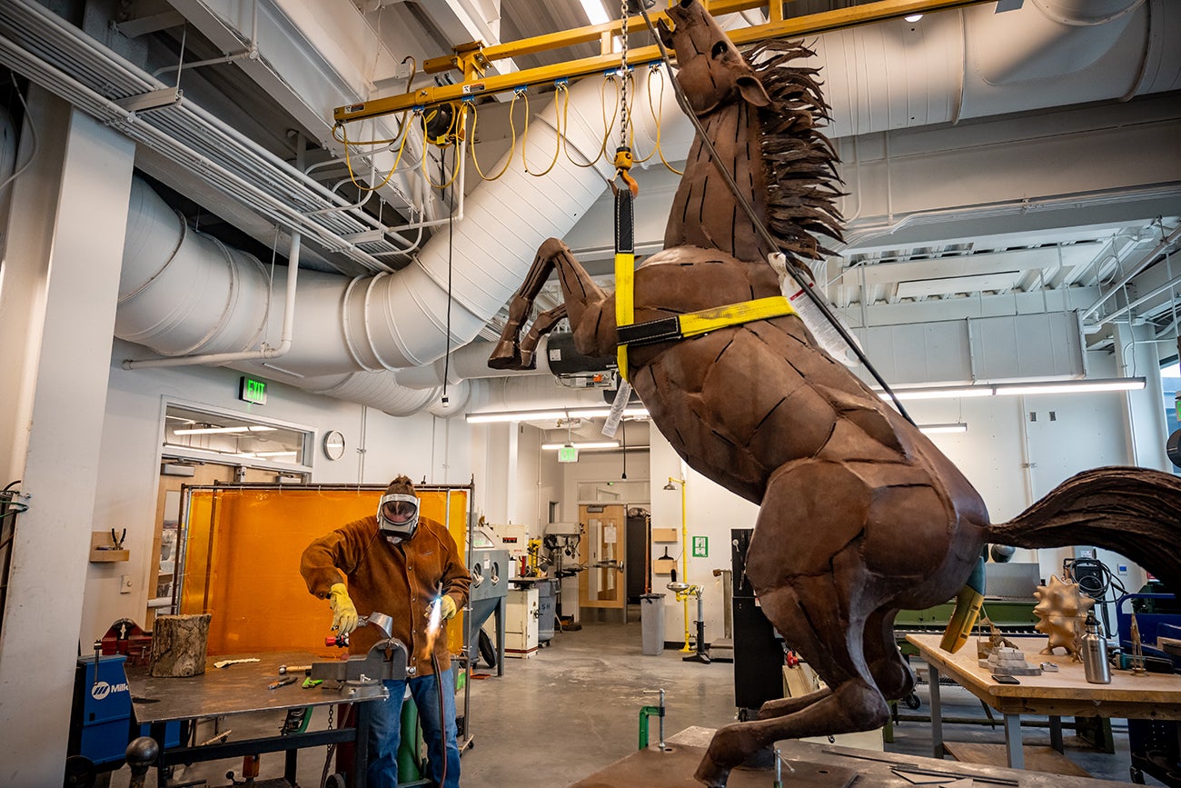 man works on piece of metal in studio, with enormous steel bronco sculpture held by harness 